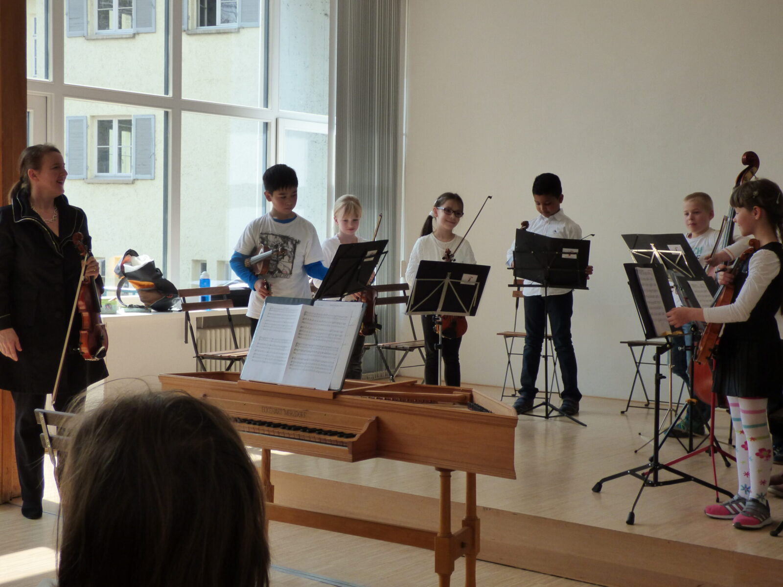 Eine Gruppe von jungen Musikern steht vor einem großen Fenster und bereitet sich auf ihre Aufführung vor. Die Kinder halten ihre Instrumente und Notenblätter, während sie sich auf ihre Darbietung konzentrieren. Im Vordergrund ist ein Holzklavier mit aufgeschlagenen Noten zu sehen, das ebenfalls Teil der bevorstehenden Aufführung ist.