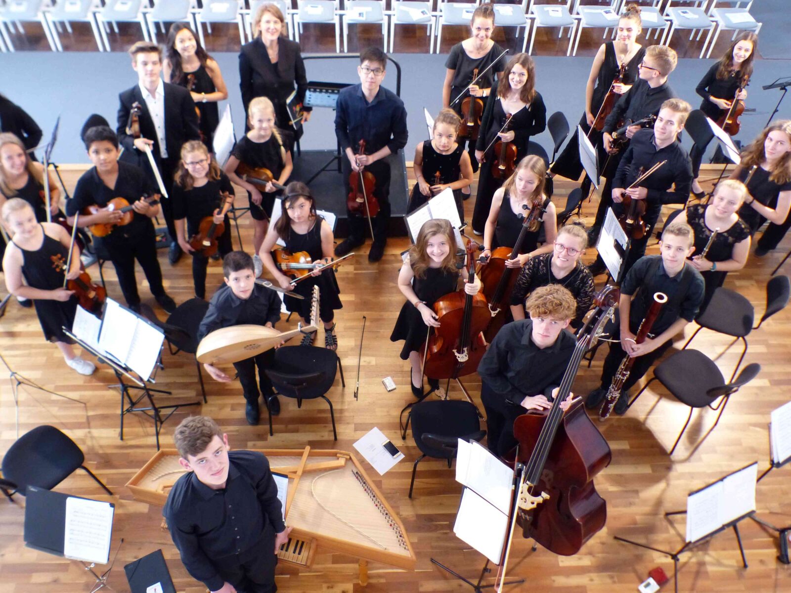 Eine Gruppe von Musikern in einem Orchester bereitet sich auf ein Konzert vor. Sie sind in einem Proberaum und tragen schwarze Kleidung. Verschiedene Instrumente wie Geigen, Celli, Kontrabässe und Blasinstrumente sind zu sehen.