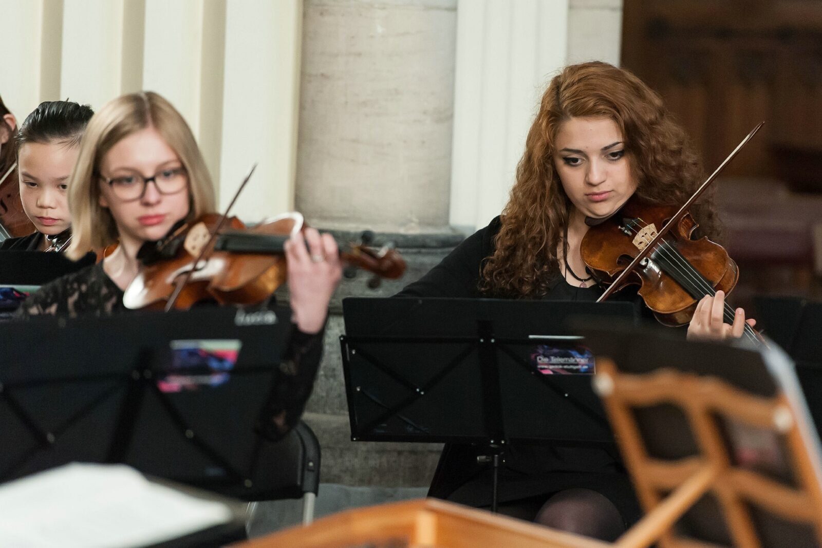 Eine Gruppe von Musikerinnen spielt Violine in einem Konzertsaal. Sie sind in schwarze Kleidung gekleidet und konzentrieren sich auf ihre Notenständer.
