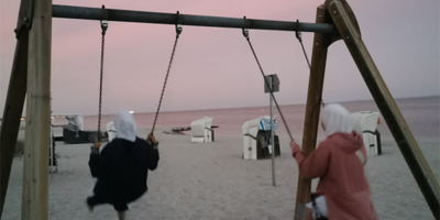 Zwei Personen schaukeln am Strand bei Sonnenuntergang. Der Himmel ist in sanften Rosa- und Lilatönen gefärbt, und im Hintergrund sind Strandkörbe sowie das Meer zu sehen. Die Szene vermittelt eine friedliche und entspannte Atmosphäre.