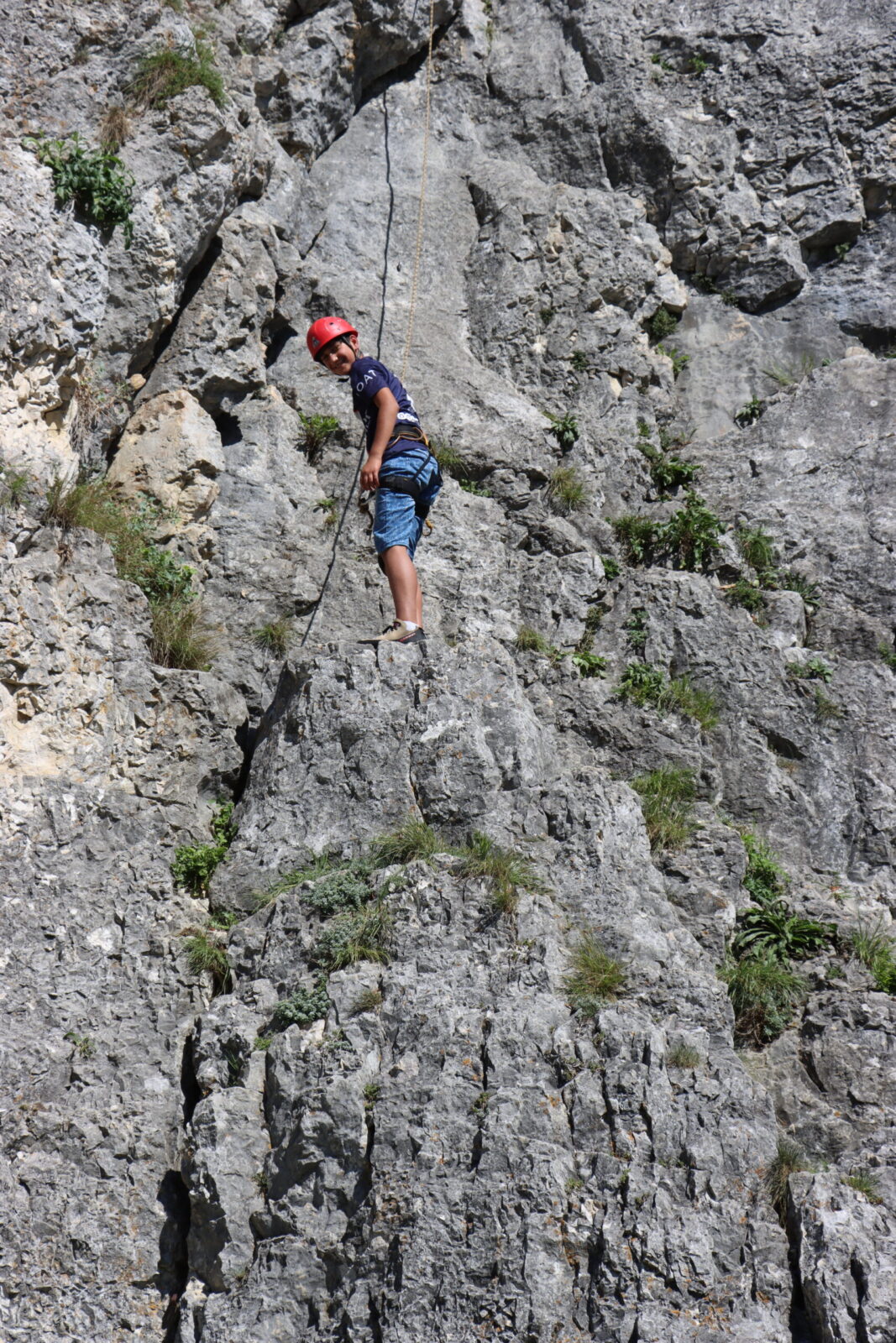 Ein Kind mit rotem Helm und Kletterausrüstung steht an einer steilen Felswand. Es trägt ein dunkelblaues T-Shirt und blaue Shorts. Das Kind ist durch ein Seil gesichert und scheint konzentriert auf den nächsten Schritt zu sein, um weiterzuklettern.