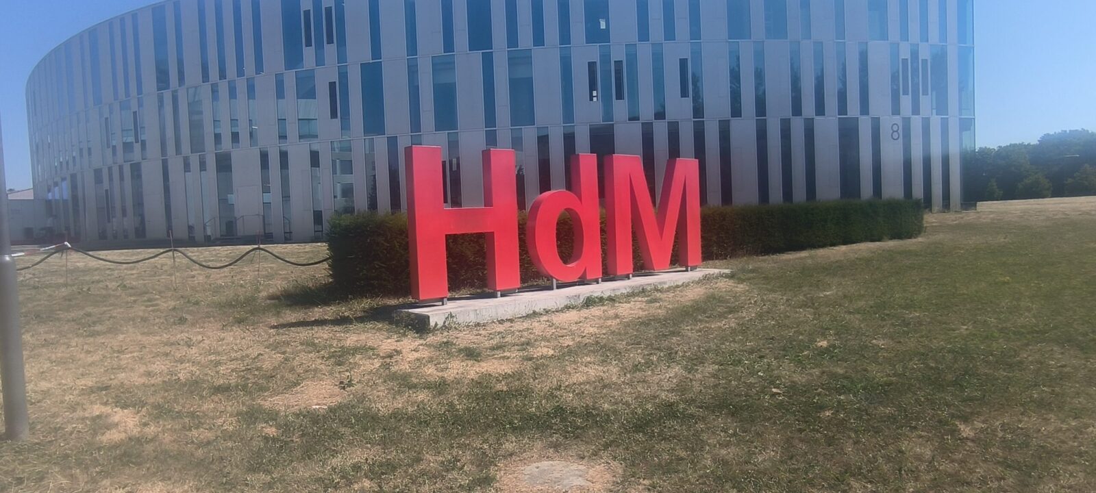 Ein großes, rotes "HdM" Schild steht vor einem modernen Gebäude mit vielen Glasfenstern. Das Schild befindet sich auf einer Betonplattform, umgeben von einer Grünfläche und Hecken. Die Sonne scheint und es herrscht klares Wetter.