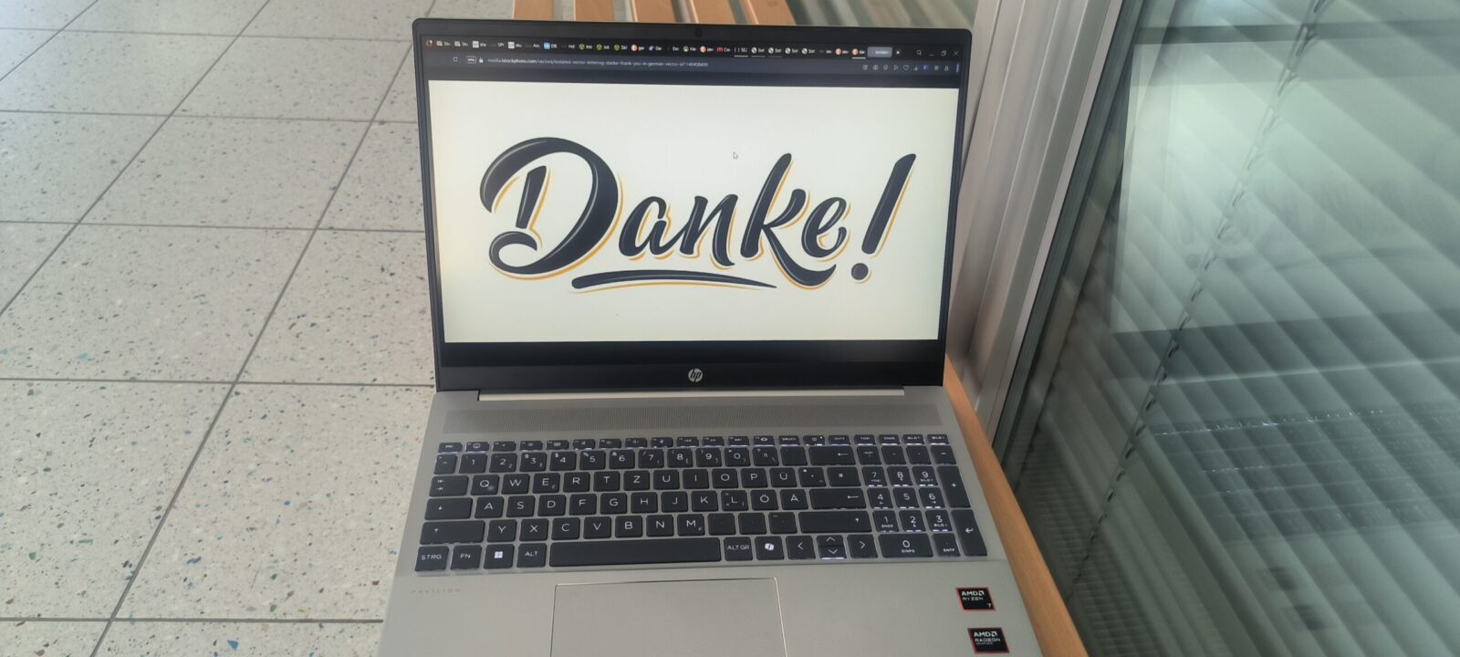 Ein silberner HP-Laptop steht auf einer Holzbank und zeigt auf seinem Bildschirm das Wort "Danke!" in stilisierter Schrift. Der Laptop befindet sich in einem modernen, hellen Raum mit Glaswänden und einem gefliesten Boden. Die Tastatur ist beleuchtet und auf dem Laptop sind zwei AMD-Aufkleber zu sehen.