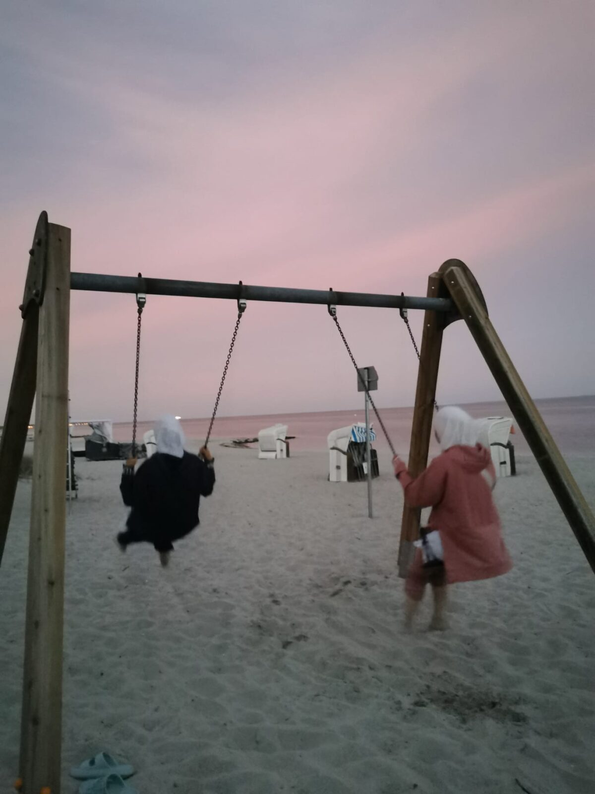 Zwei Personen schaukeln am Strand bei Sonnenuntergang. Der Himmel ist in sanften Rosa- und Lilatönen gefärbt, und im Hintergrund sind Strandkörbe sowie das Meer zu sehen. Die Szene vermittelt eine friedliche und entspannte Atmosphäre.