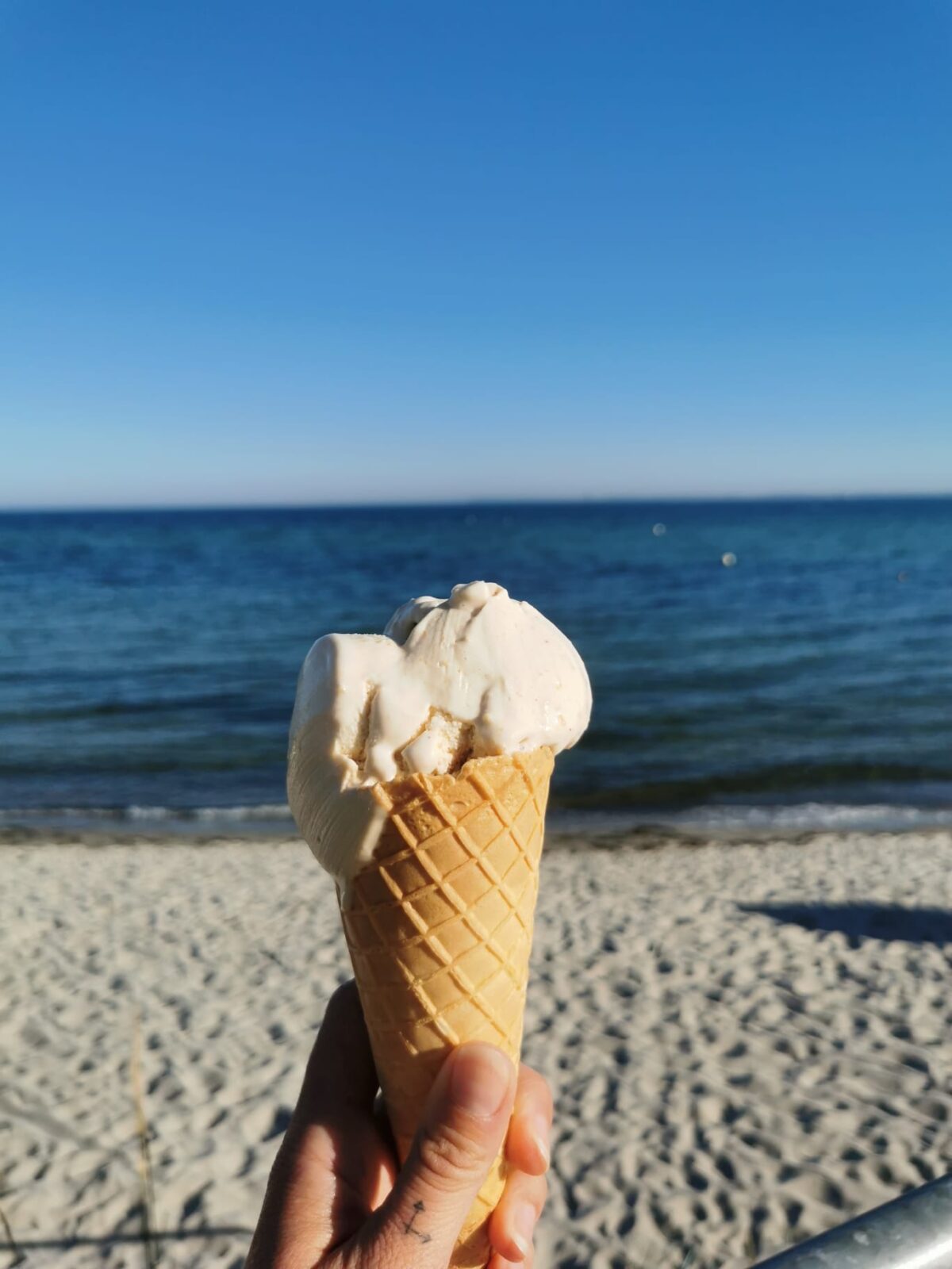 Eine Hand hält ein Eis in einer Waffel vor einem sandigen Strand mit blauem Meer im Hintergrund. Der Himmel ist klar und blau, und die Szene vermittelt ein Gefühl von Sommer und Erholung. Das Eis scheint Vanille oder eine ähnliche helle Geschmacksrichtung zu sein.