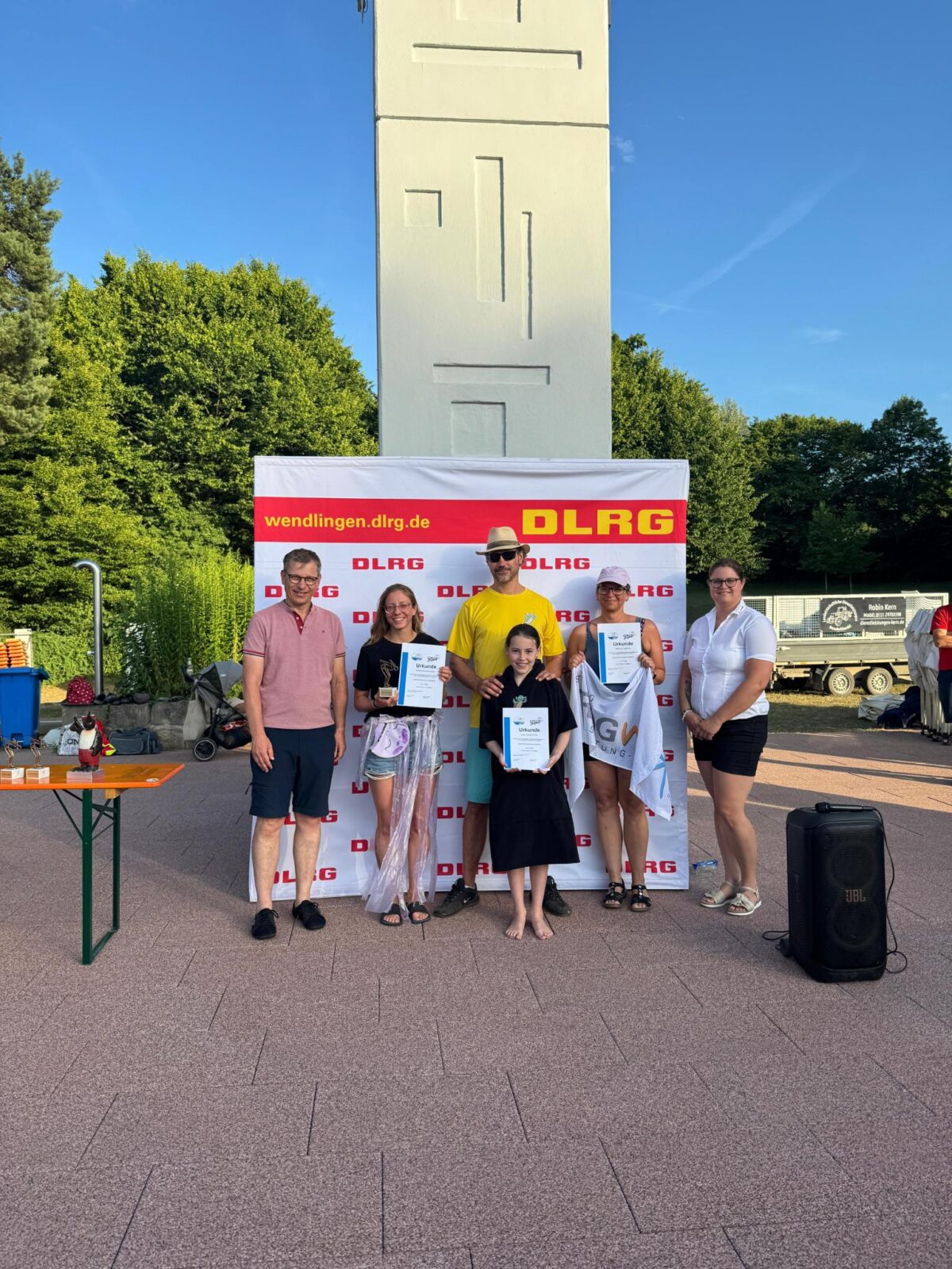 Eine Gruppe von Menschen steht vor einem DLRG-Banner und hält Urkunden in den Händen. Im Hintergrund sind grüne Bäume und ein weißer Turm zu sehen. Ein Tisch mit Pokalen und eine Lautsprecherbox sind ebenfalls im Bild.