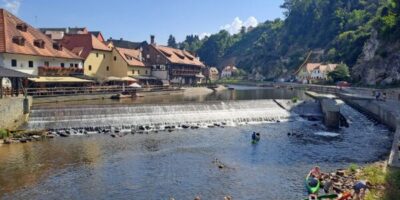 Ein malerisches Bild eines Flusses mit einem kleinen Wasserfall, umgeben von traditionellen Gebäuden mit roten Dächern und Fachwerk. Im Vordergrund paddeln Menschen in Kanus auf dem ruhigen Wasser. Im Hintergrund sind bewaldete Hügel und ein klarer blauer Himmel zu sehen.
