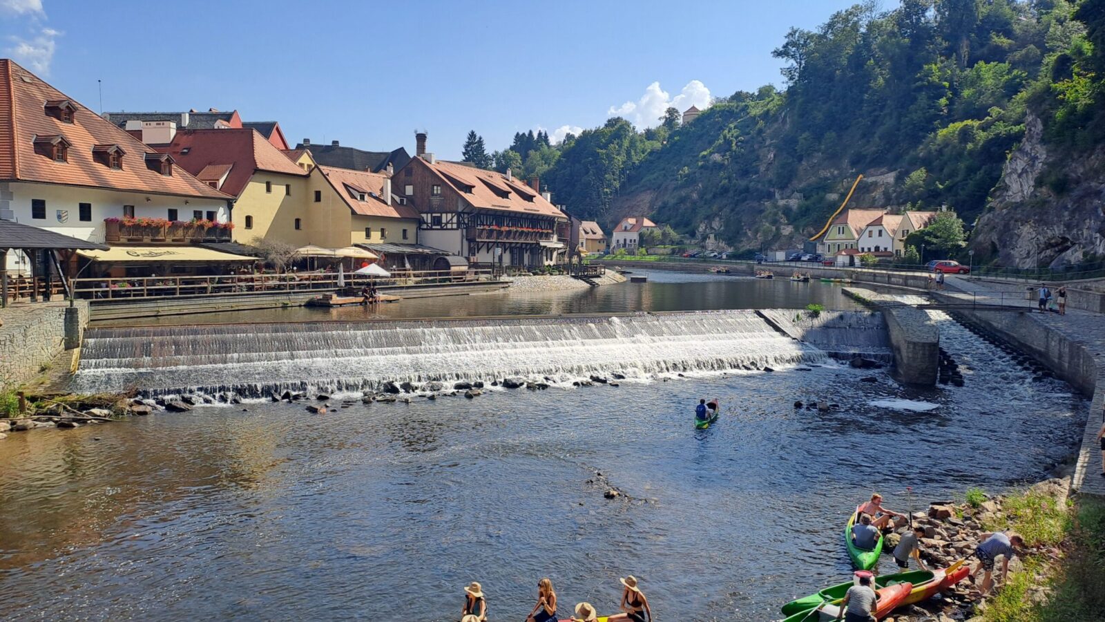 Ein malerisches Bild eines Flusses mit einem kleinen Wasserfall, umgeben von traditionellen Gebäuden mit roten Dächern und Fachwerk. Im Vordergrund paddeln Menschen in Kanus auf dem ruhigen Wasser. Im Hintergrund sind bewaldete Hügel und ein klarer blauer Himmel zu sehen.