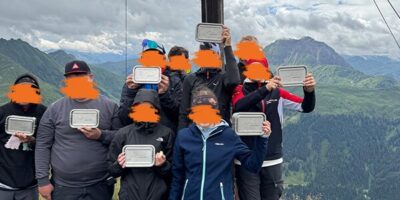 Eine Gruppe von Wanderern steht auf einem Berggipfel neben einem großen Holzkreuz und hält Brotzeitboxen in den Händen. Im Hintergrund erstreckt sich eine malerische Berglandschaft unter einem bewölkten Himmel. Die Wanderer sind warm angezogen und tragen Wanderschuhe.