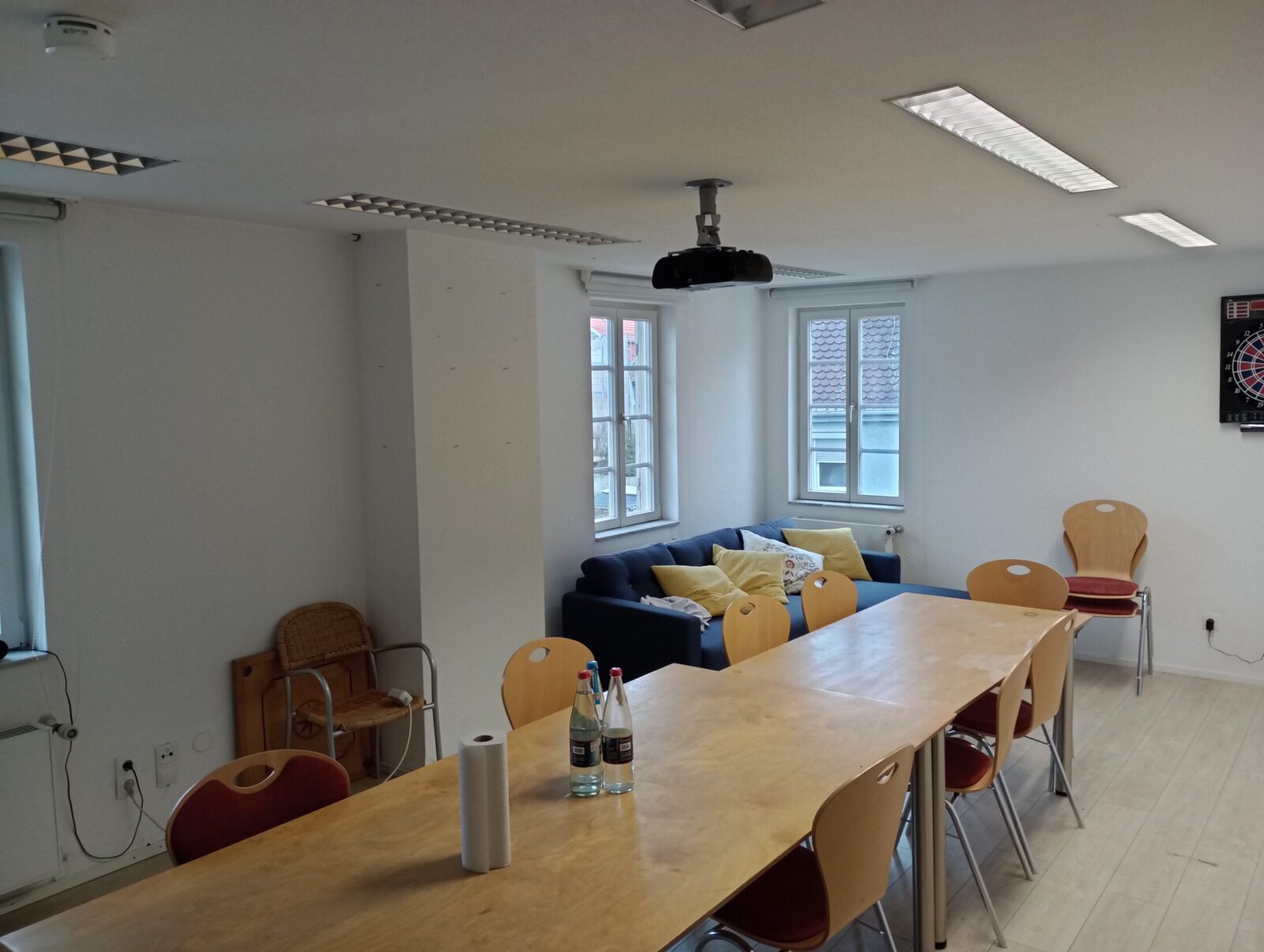 Ein Besprechungsraum mit einem langen Holztisch und mehreren Holzstühlen. Im Hintergrund befindet sich eine blaue Couch mit gelben Kissen, die vor zwei Fenstern steht. An der Decke ist ein Projektor montiert und auf dem Tisch stehen Wasserflaschen und eine Rolle Küchenpapier.