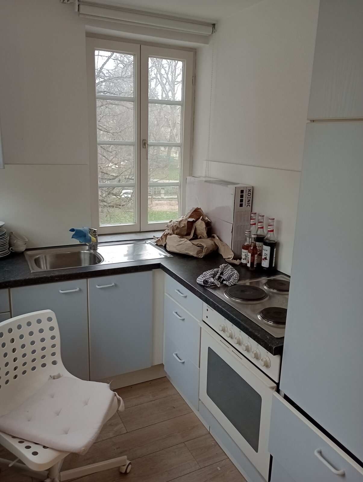 Eine kleine Küche mit weiß-blauen Schränken und einem Fenster, das einen Blick auf einen bewaldeten Bereich bietet. Auf der Arbeitsplatte liegen Papiertüten, einige Flaschen und ein Geschirrtuch. Ein weißer Stuhl mit Kissen steht vor der Spüle und der Herd ist auf der rechten Seite zu sehen.