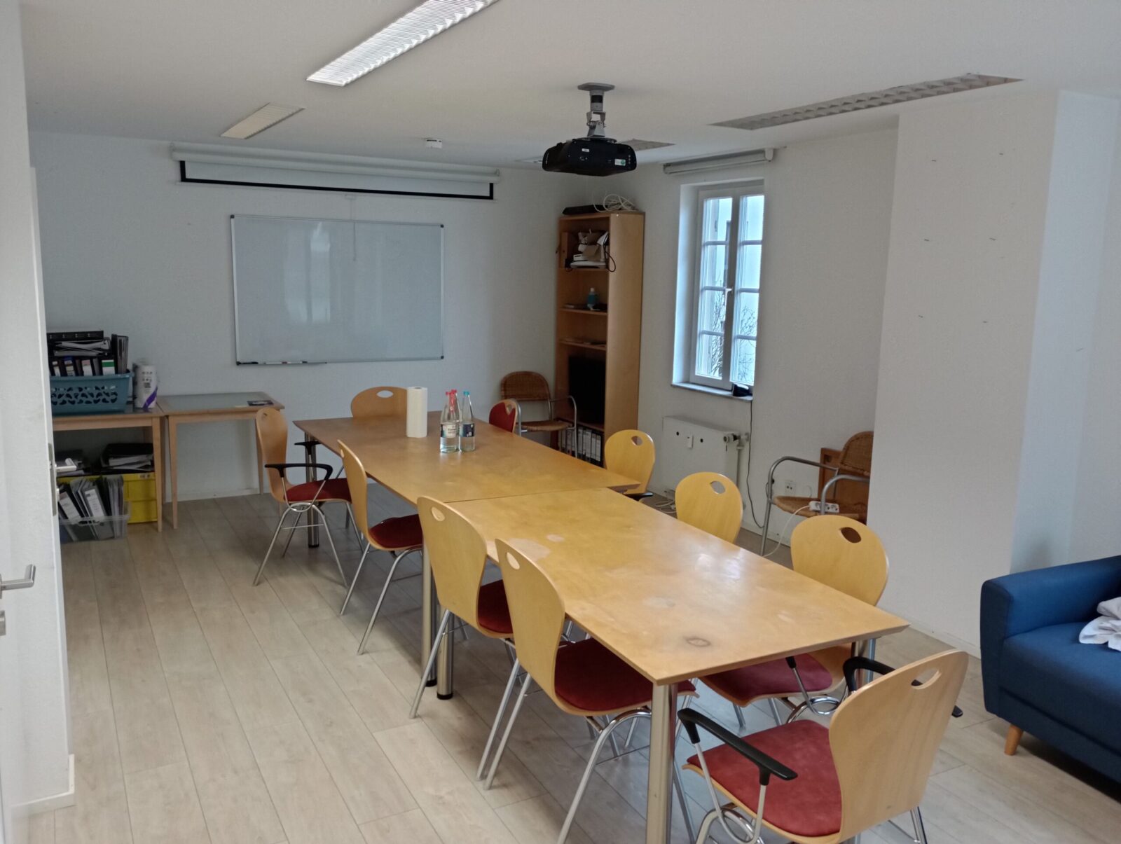 Ein Konferenzraum mit einem langen Holztisch, umgeben von roten und gelben Stühlen. An der Decke hängt ein Projektor, und im Hintergrund ist eine weiße Tafel sowie ein Regal mit Büchern und Ordnern zu sehen. Zwei Wasserflaschen stehen auf dem Tisch, und durch die Fenster fällt Tageslicht in den Raum.