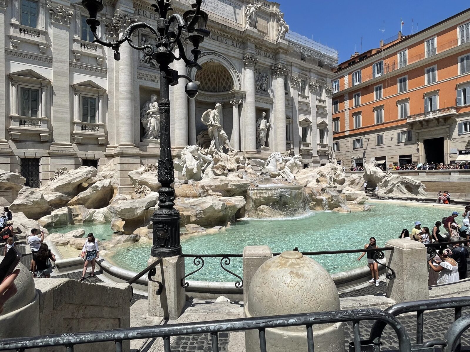 Das Bild zeigt den berühmten Trevi-Brunnen in Rom, Italien, an einem sonnigen Tag. Im Vordergrund ist ein dekorativer Laternenpfahl und ein Teil des Brunnens mit klar blauem Wasser zu sehen. Im Hintergrund sind beeindruckende barocke Skulpturen und die Fassade des Gebäudes sichtbar.