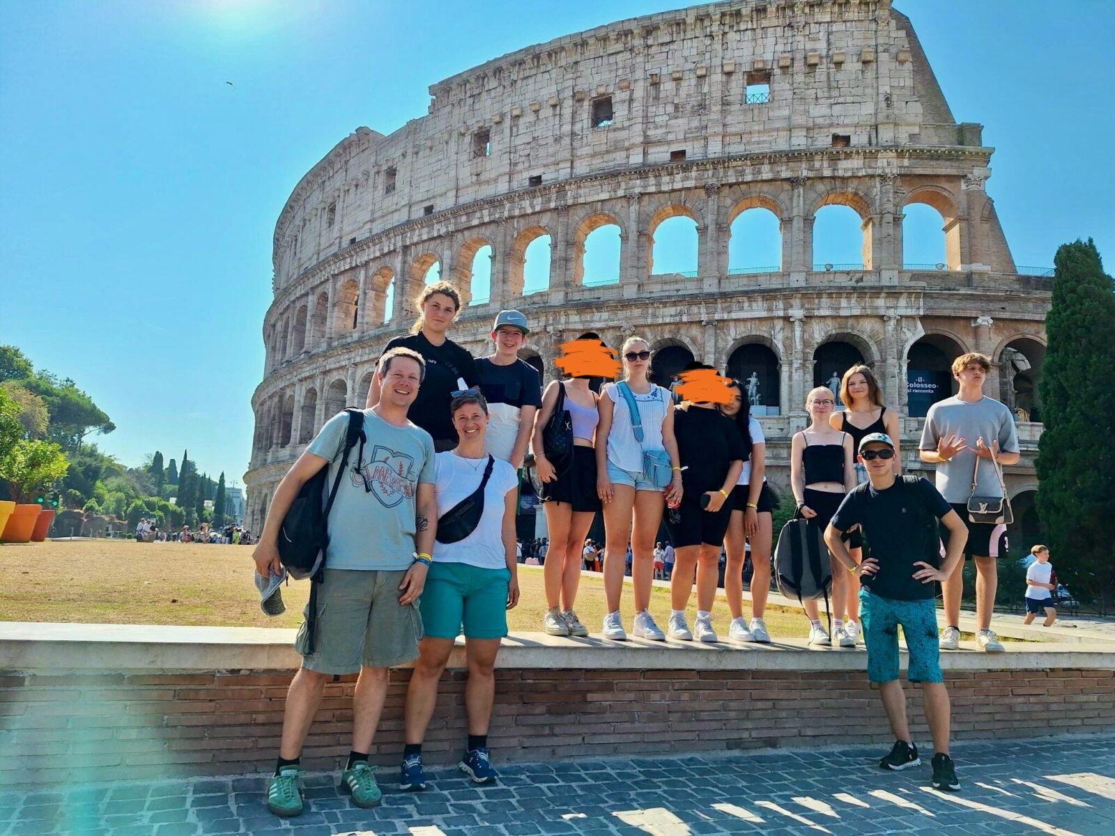 Eine Gruppe von Menschen steht vor dem Kolosseum in Rom, Italien. Das historische Amphitheater erstreckt sich majestätisch im Hintergrund. Die Personen tragen sommerliche Kleidung und stehen lächelnd nebeneinander.