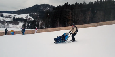 Eine Frau fährt mit Skiern einen schneebedeckten Hang hinunter und hat vor sich ein Skibob, welches Sie führt. Sie und der Junge auf dem Skibob tragen einen Helm und dicke Winterkleidung. Im Hintergrund sind weitere Personen zu sehen, die an einer Holzbarriere stehen, sowie eine bewaldete Landschaft und schneebedeckte Hügel.
