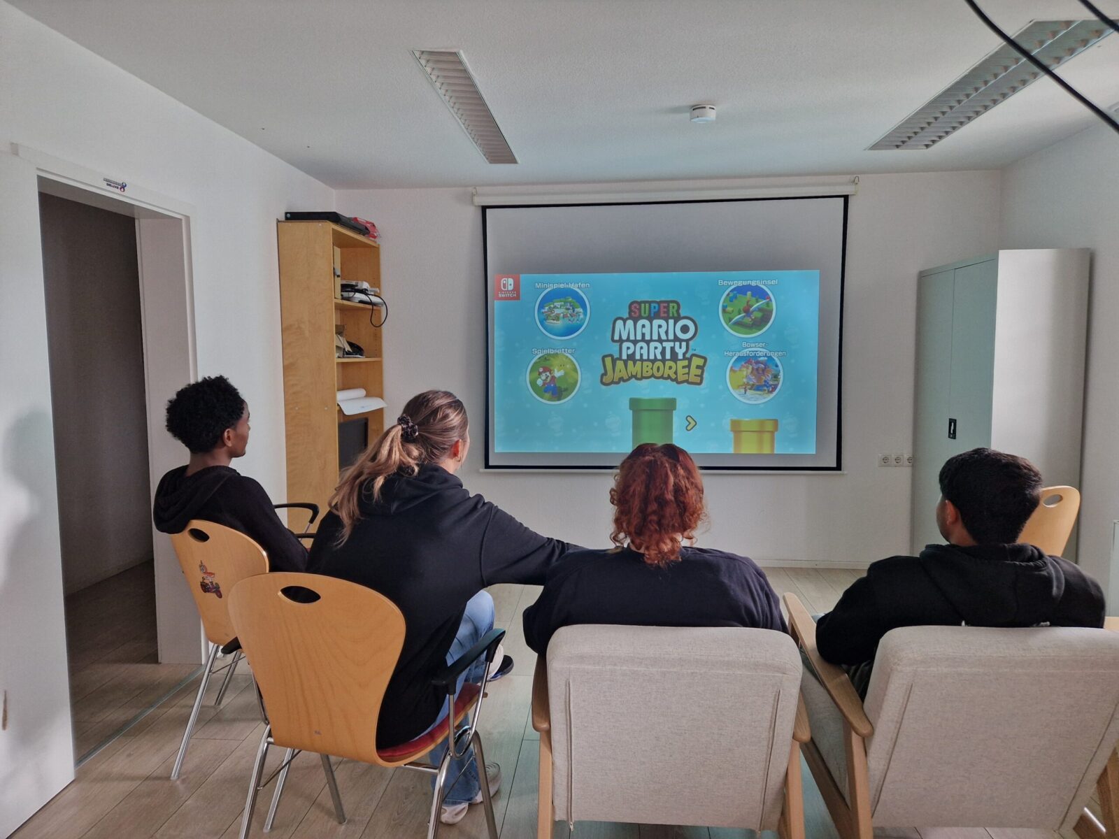 Vier Personen sitzen in einem Raum auf Stühlen und schauen auf eine Leinwand, auf der das Spiel "Super Mario Party Jamboree" von Nintendo angezeigt wird. Die Atmosphäre wirkt entspannt, und die Gruppe scheint sich auf eine gemeinsame Spielrunde vorzubereiten. Im Hintergrund sind ein Regal und eine Tür zu sehen, was auf eine informelle Umgebung hinweist.