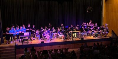 Eine Musikgruppe und ein Chor treten gemeinsam auf einer Bühne in einem Saal auf. Die Bühne ist mit einer schwarzen Rückwand und Bühnenbeleuchtung ausgestattet, während das Publikum in Reihen davor sitzt. Verschiedene Instrumente wie Blasinstrumente und ein Klavier sind zu sehen, und die Atmosphäre wirkt lebendig und konzertartig.
