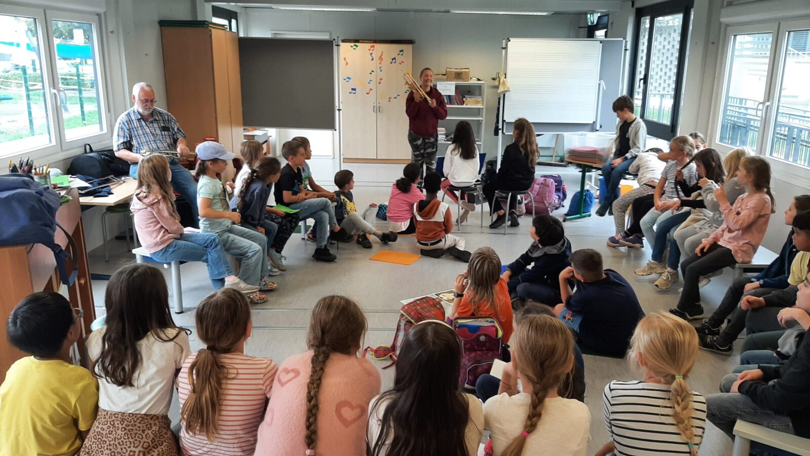Eine Gruppe von Kindern sitzt in einem Halbkreis in einem Klassenzimmer und hört einer Person zu, die ein Musikinstrument präsentiert. Der Raum ist hell und freundlich gestaltet, mit großen Fenstern, einem Whiteboard und Schränken, die mit bunten Noten dekoriert sind. Im Hintergrund sind Schultaschen und Materialien zu sehen, die eine lebendige Lernatmosphäre vermitteln.
