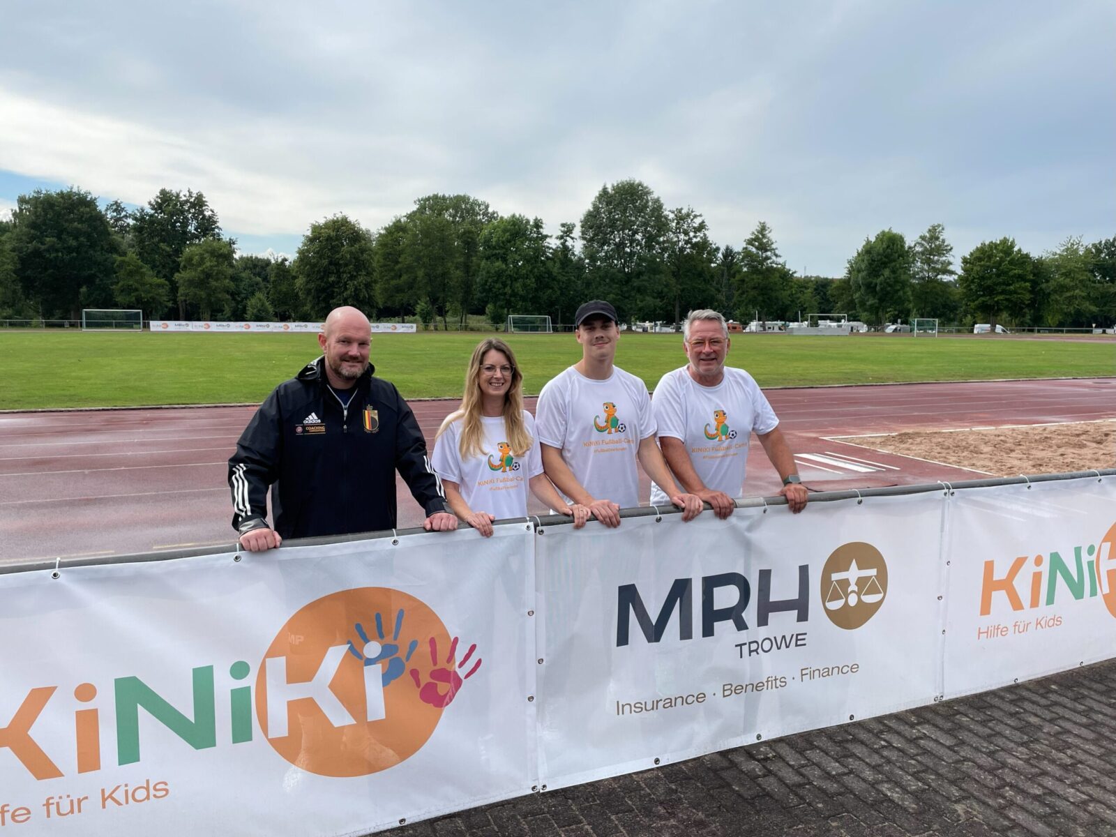 Vier Personen stehen hinter einem Werbebanner mit den Logos von "KiNiKi – Hilfe für Kids" und "MRH Trowe – Insurance, Benefits, Finance" vor einer Sportanlage mit Laufbahn und Rasenfläche. Im Hintergrund sind Bäume und ein bewölkter Himmel zu sehen. Die Personen tragen Sportkleidung und T-Shirts mit dem Logo von KiNiKi.