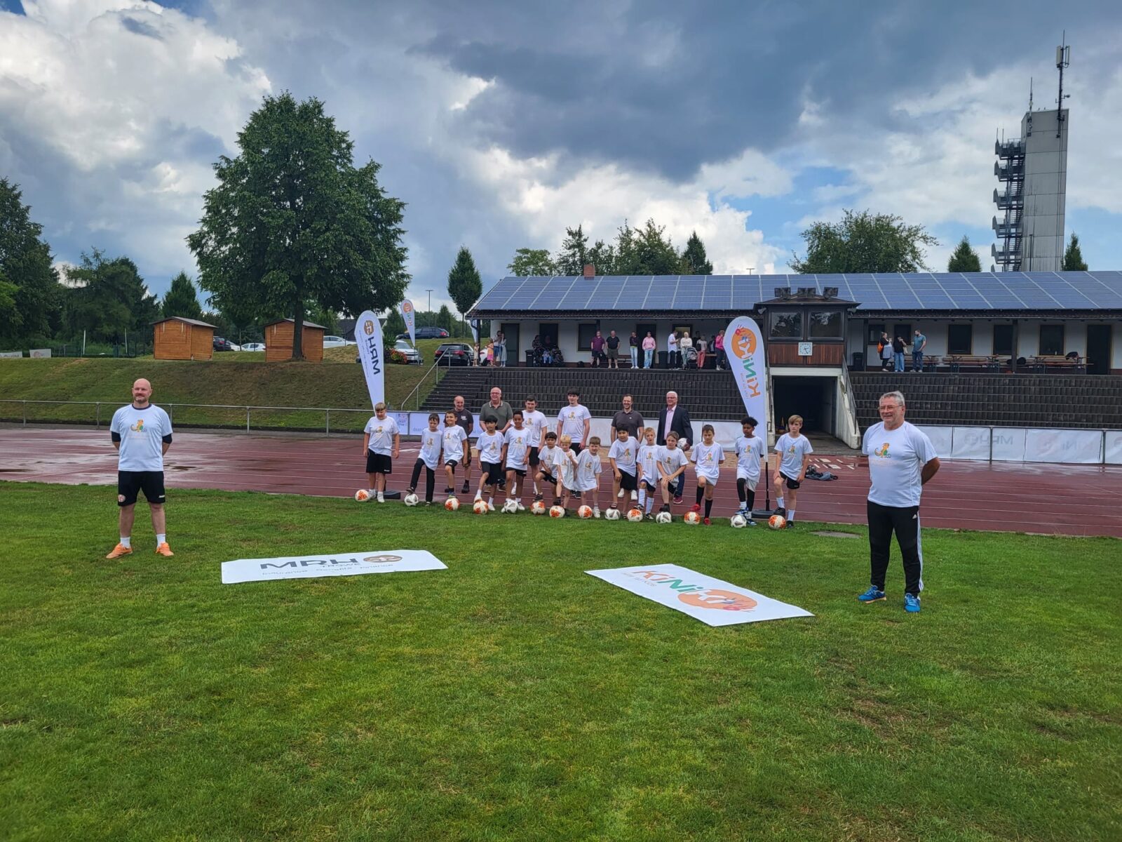 Eine Gruppe von Kindern und Erwachsenen posiert auf einem Sportplatz für ein Gruppenfoto. Im Vordergrund sind zwei Banner mit Logos von Sponsoren zu sehen, während im Hintergrund ein Gebäude mit Solarpanelen und Zuschauer auf einer Tribüne sichtbar sind. Die Teilnehmer tragen weiße T-Shirts und sind von Fußbällen umgeben, was auf ein Fußball-Event hindeutet.