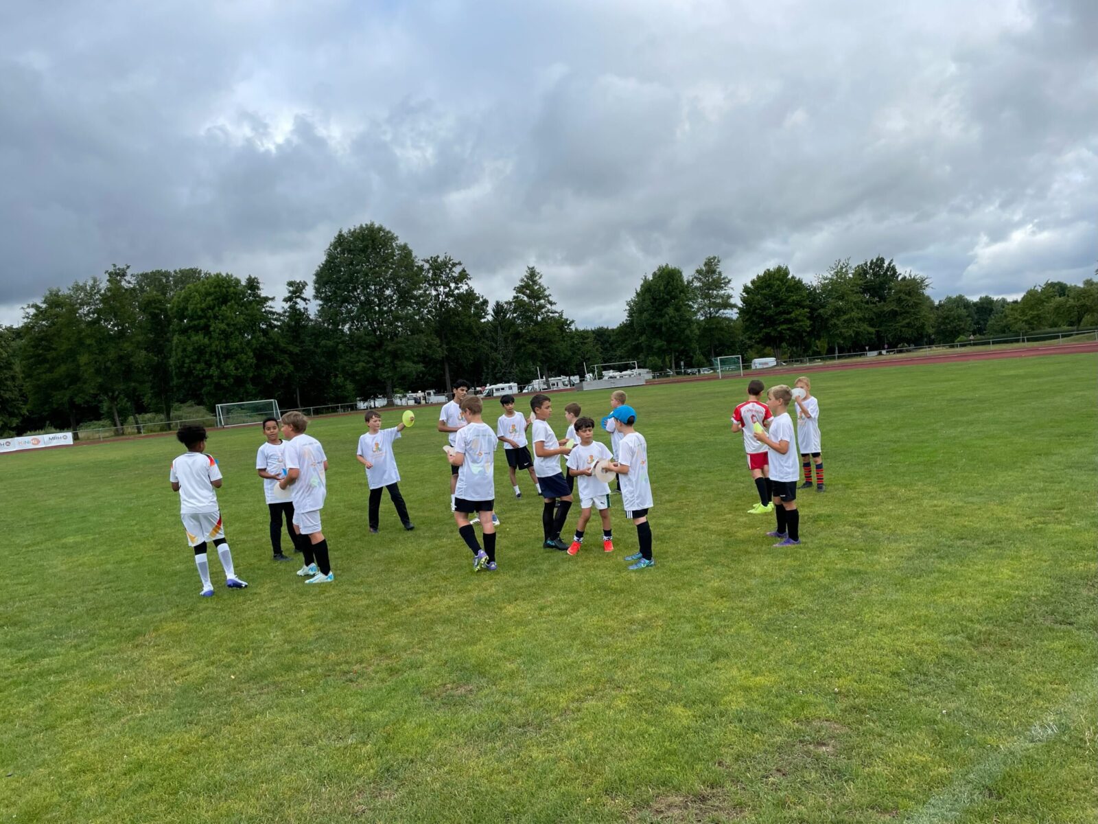Eine Gruppe von Kindern in weißen Trikots steht auf einem grünen Fußballfeld und unterhält sich oder spielt miteinander. Im Hintergrund sind Bäume, Fußballtore und eine Laufbahn zu sehen, während der Himmel bewölkt ist. Die Szene vermittelt eine entspannte und sportliche Atmosphäre.