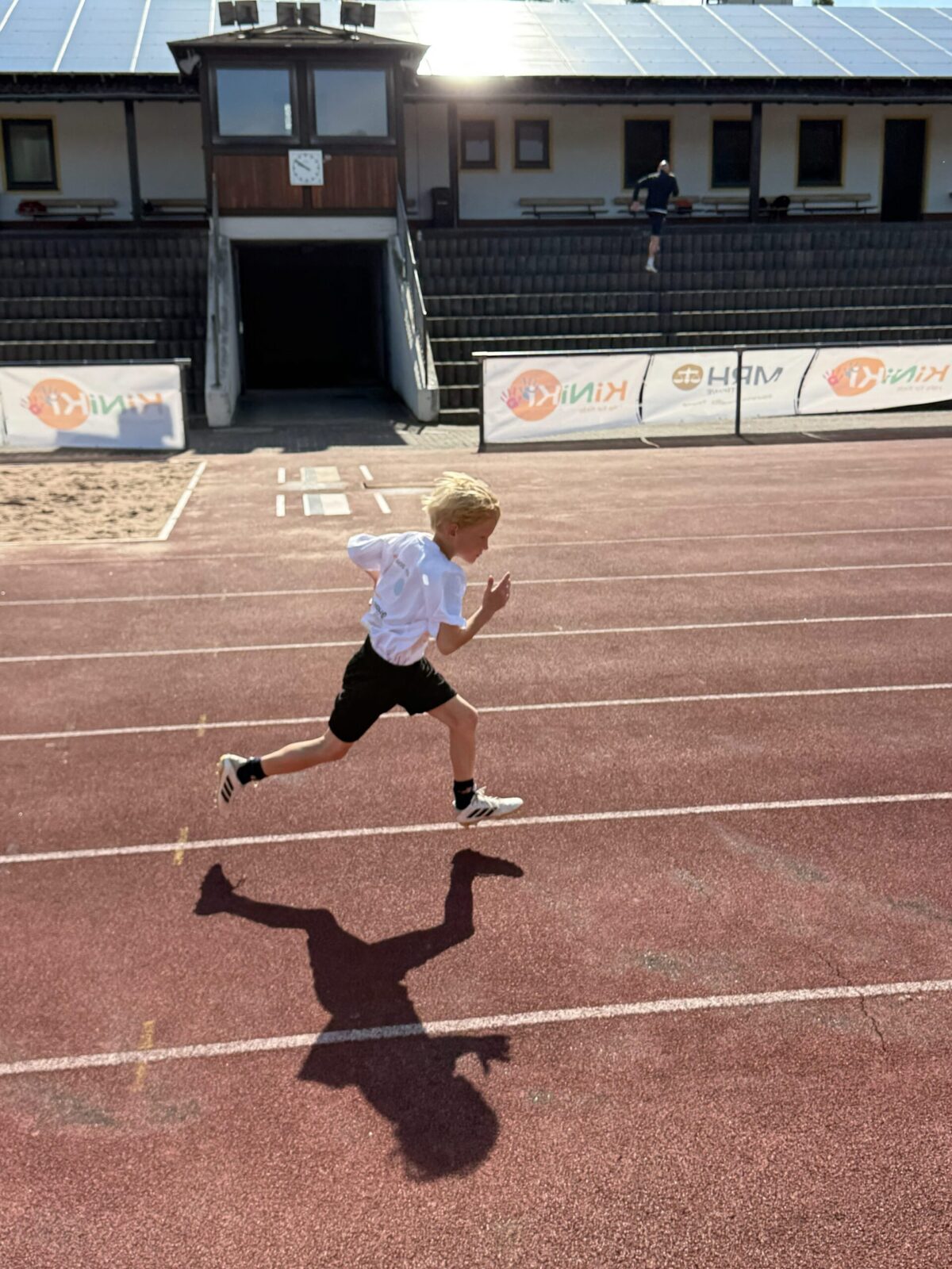 Ein Kind in sportlicher Kleidung läuft auf einer roten Laufbahn eines Stadions, während die Sonne einen deutlichen Schatten auf den Boden wirft. Im Hintergrund sind eine Tribüne mit Sitzplätzen, ein Gebäude mit einer Uhr sowie Werbebanner zu sehen. Die Szene vermittelt Dynamik und Energie in einer sportlichen Umgebung.