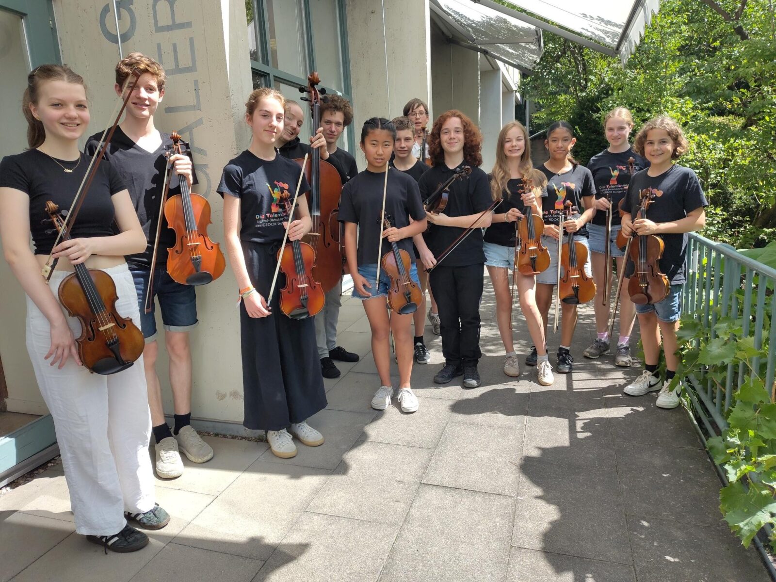 Eine Gruppe junger Musikerinnen und Musiker steht mit ihren Streichinstrumenten, darunter Geigen und Bratschen, auf einer sonnigen Terrasse. Sie tragen einheitliche schwarze T-Shirts mit einem bunten Logo und sind umgeben von grüner Natur und einem Gebäude im Hintergrund. Die Stimmung wirkt konzentriert und harmonisch, passend zu einem musikalischen Anlass.