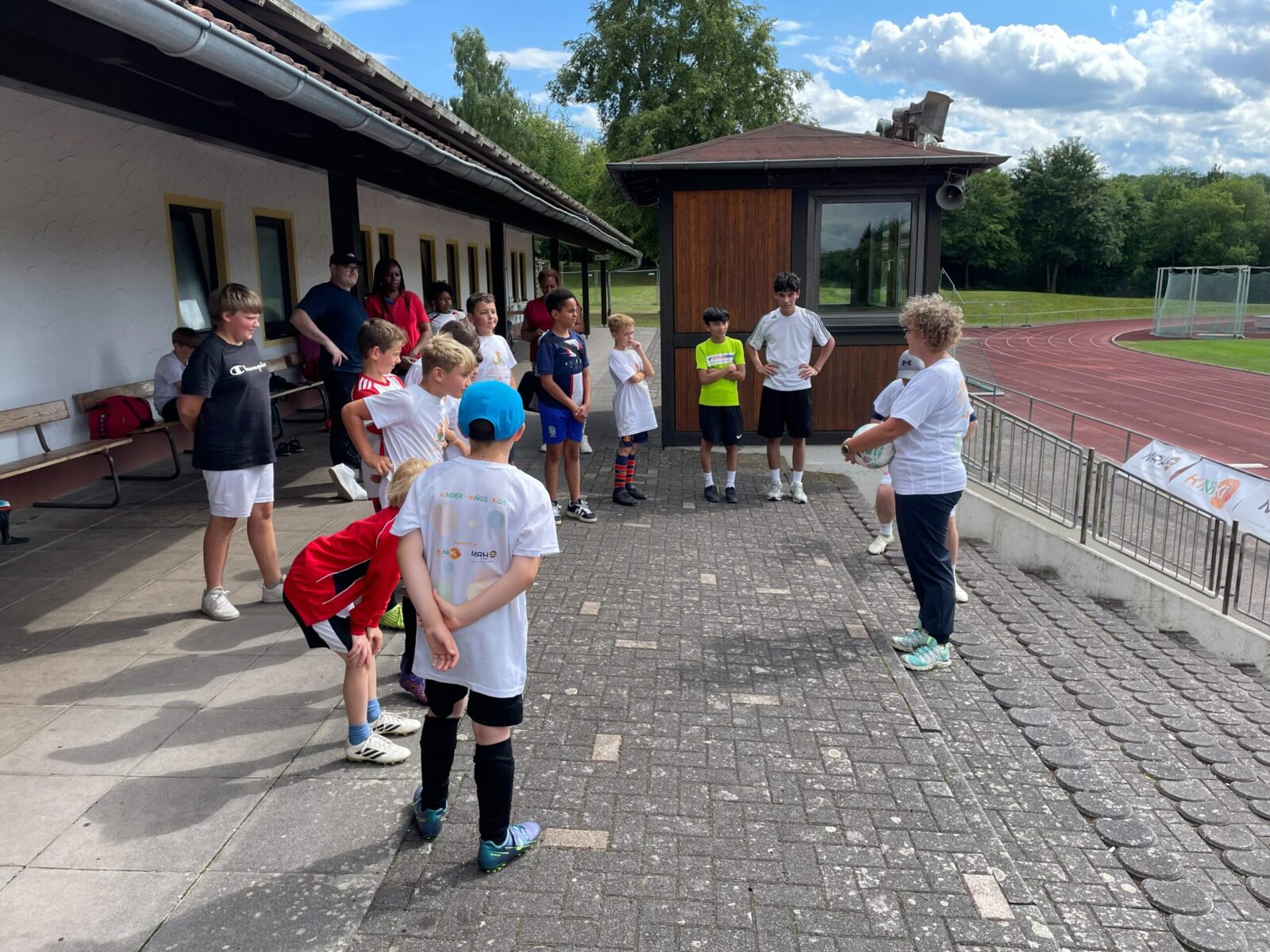 Eine Gruppe von Kindern und Jugendlichen steht in sportlicher Kleidung auf einem gepflasterten Platz vor einem Gebäude mit gelben Fenstern. Eine erwachsene Person, die einen Ball hält, spricht zu der Gruppe, während im Hintergrund eine Laufbahn und grüne Bäume zu sehen sind. Der Himmel ist blau mit einigen Wolken, was auf einen sonnigen Tag hinweist.