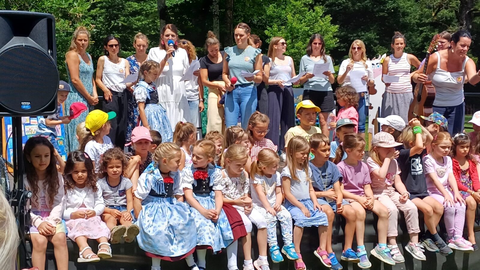 Eine Gruppe von Kindern sitzt auf einer Bank im Freien, einige tragen traditionelle Trachtenkleidung. Im Hintergrund stehen Erwachsene, einige mit Mikrofonen und Notizen, während sie offenbar eine Veranstaltung moderieren oder begleiten. Die Szene spielt sich an einem sonnigen Tag in einer grünen, bewaldeten Umgebung ab.