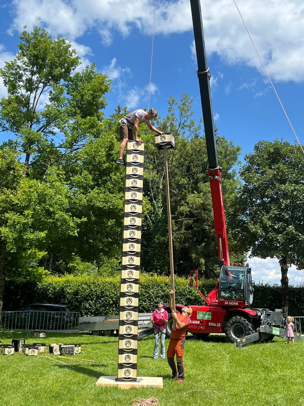 Ein Mann balanciert auf einem hohen Turm aus gestapelten Getränkekisten, während er eine weitere Kiste oben hinzufügt. Eine Person am Boden sichert den Turm mit einem Seil, während im Hintergrund ein roter Kran und grüne Bäume zu sehen sind. Der sonnige Tag und die Wiese schaffen eine lebhafte Atmosphäre für diese Outdoor-Aktivität.
