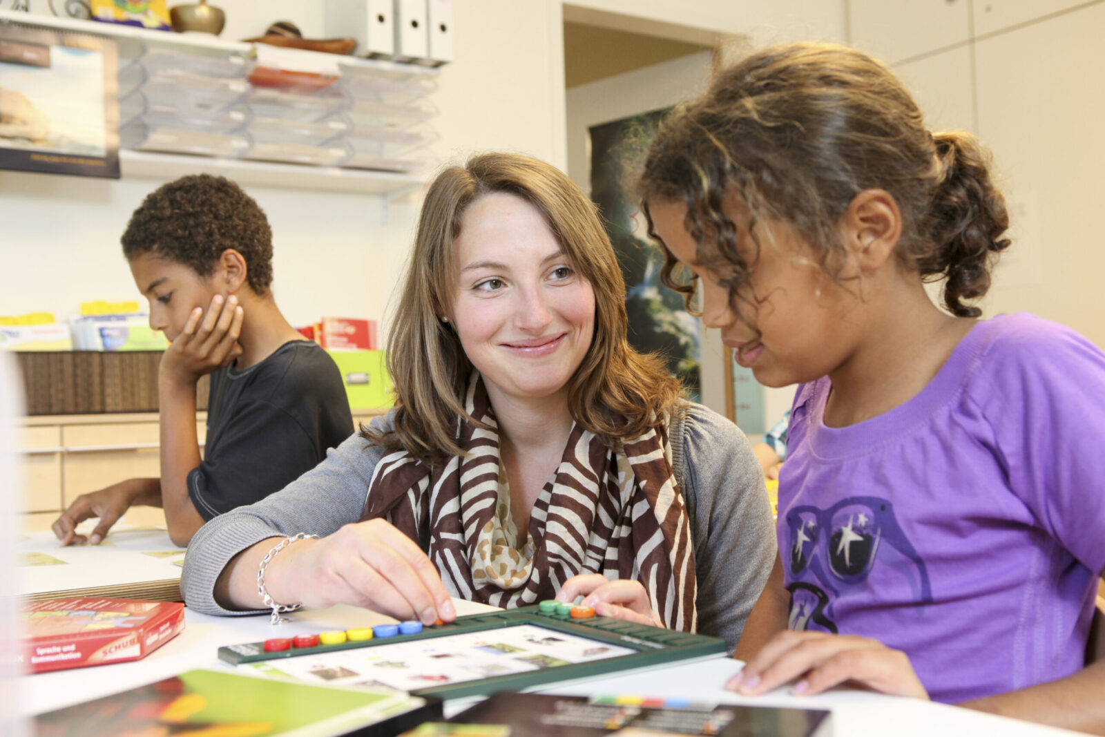 Eine Frau und ein Kind sitzen gemeinsam an einem Tisch und arbeiten an einem Lernspiel oder einer Aufgabe mit bunten Knöpfen und Bildern. Die Szene spielt in einem hellen Raum, der wie ein Klassenzimmer oder eine pädagogische Einrichtung aussieht. Die Frau trägt einen gemusterten Schal, während das Kind ein lila T-Shirt mit einem Sternenmuster trägt.