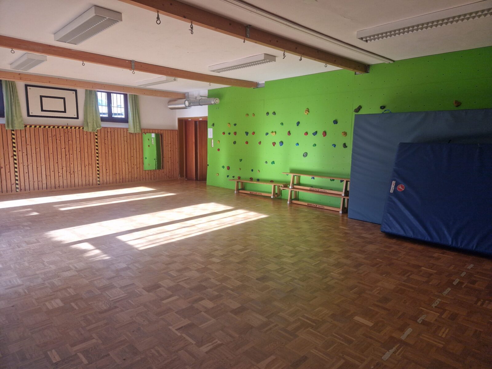 Ein heller Sportraum mit Holzboden und einer grünen Kletterwand, die mit bunten Griffen ausgestattet ist. Im Raum befinden sich Turnbänke und Matten, die an der Wand lehnen. Sonnenlicht fällt durch die Fenster und erzeugt eine freundliche Atmosphäre.