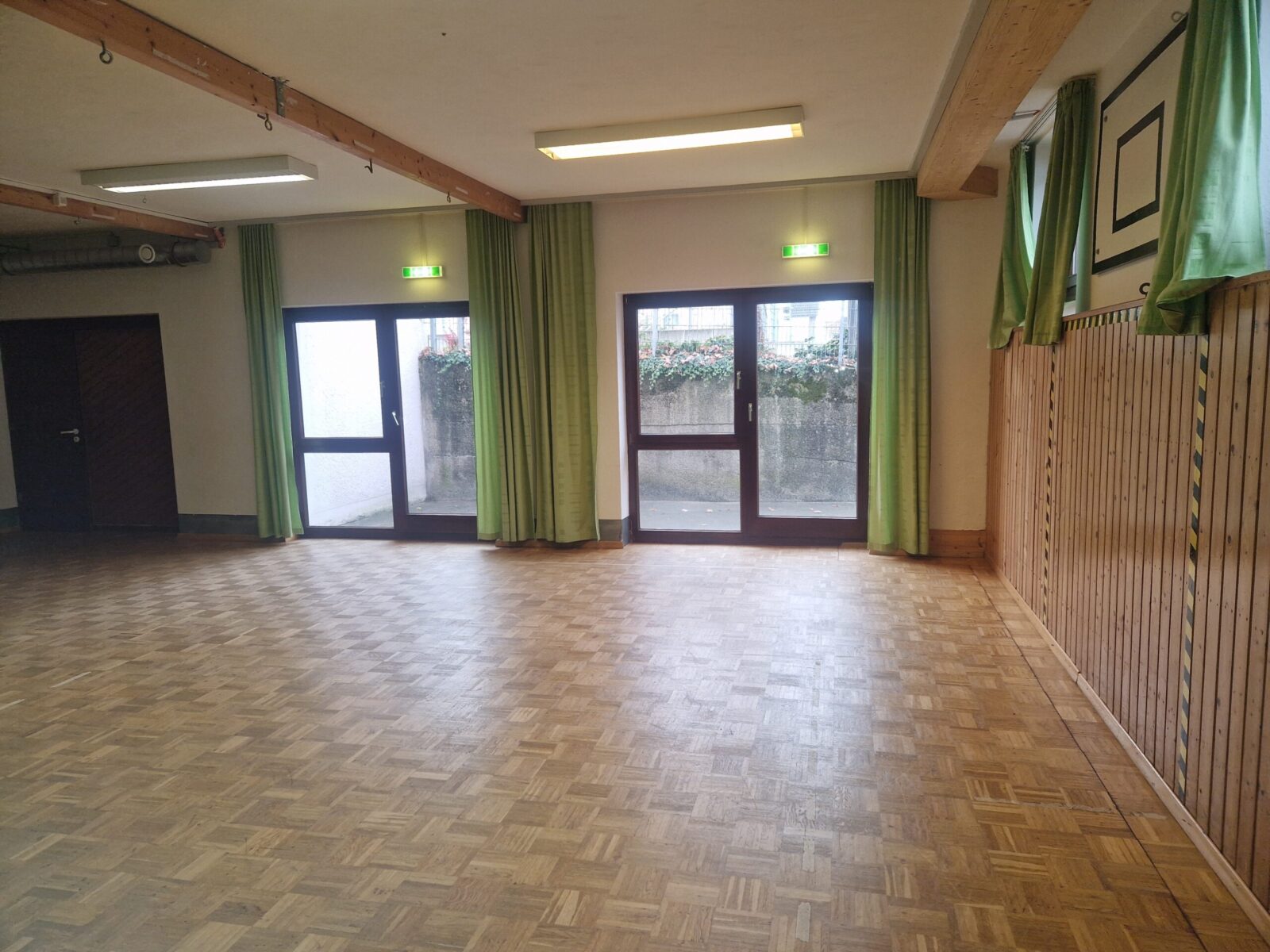 Ein leerer Raum mit Parkettboden und zwei großen Glastüren, die nach draußen führen. Die Türen sind von grünen Vorhängen eingerahmt, und oberhalb der Türen befinden sich beleuchtete Notausgangsschilder. Der Raum wirkt hell durch die natürliche Beleuchtung und die weißen Wände.