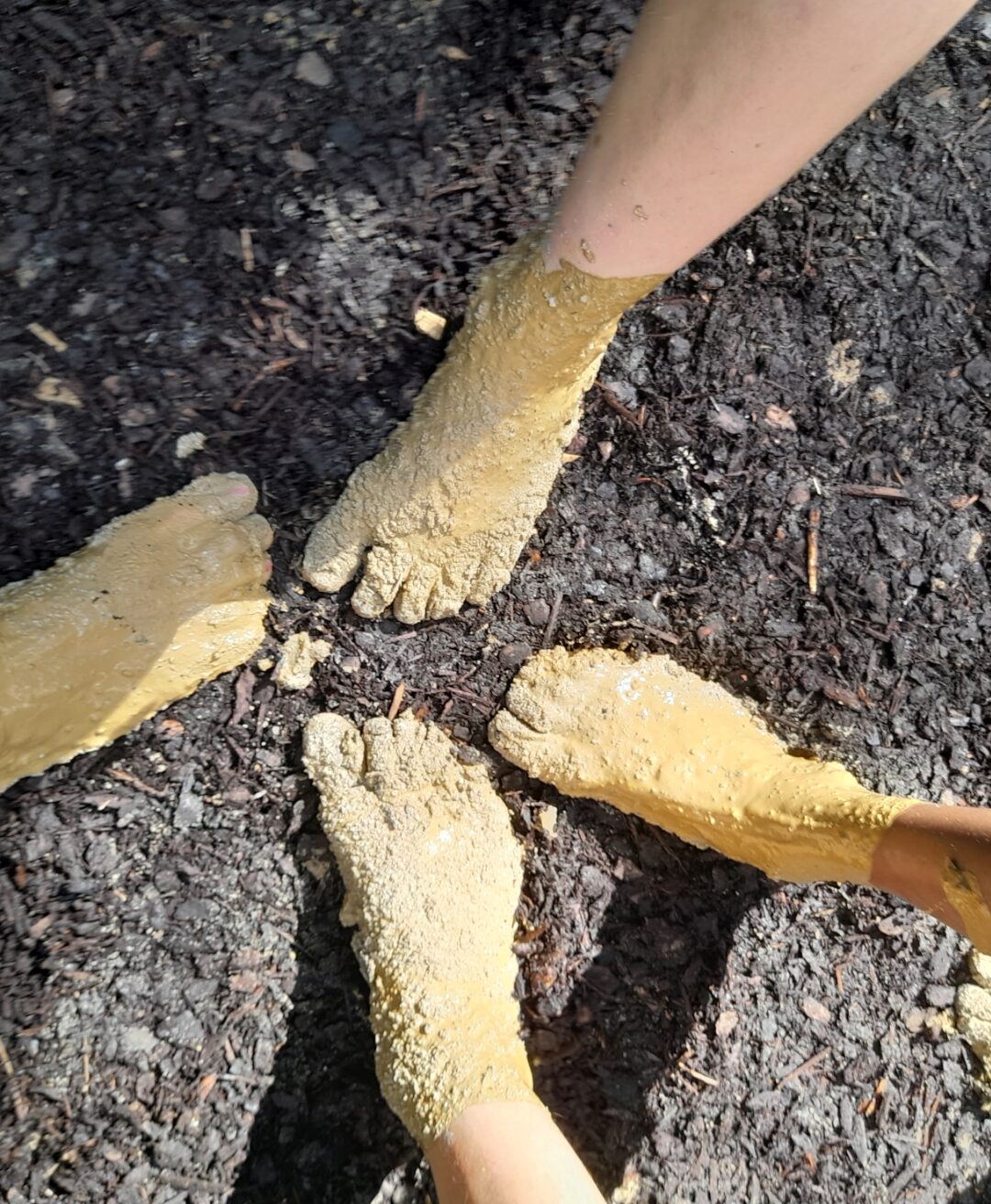 Drei Paar Füße, die mit gelbem Schlamm bedeckt sind, stehen auf einem dunklen, erdigen Untergrund. Die Füße sind barfuß und der Schlamm scheint frisch aufgetragen zu sein, was auf eine Outdoor-Aktivität oder eine Schlammanwendung hindeutet. Die Szene vermittelt ein natürliches und erdverbundenes Gefühl.