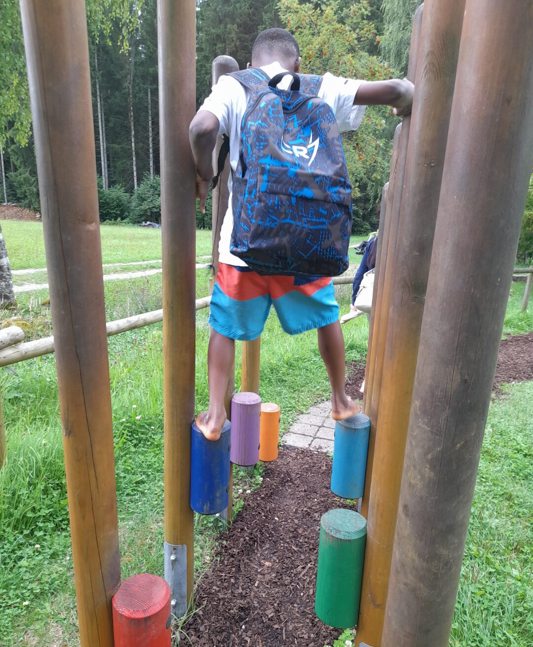 Ein Kind mit einem blauen Rucksack balanciert barfuß auf bunten Holzpfosten eines Kletterparcours im Freien. Im Hintergrund ist eine grüne Wiese sowie ein Wald zu sehen, was auf eine naturnahe Umgebung hinweist. Die Szene vermittelt einen aktiven Moment in der Natur.