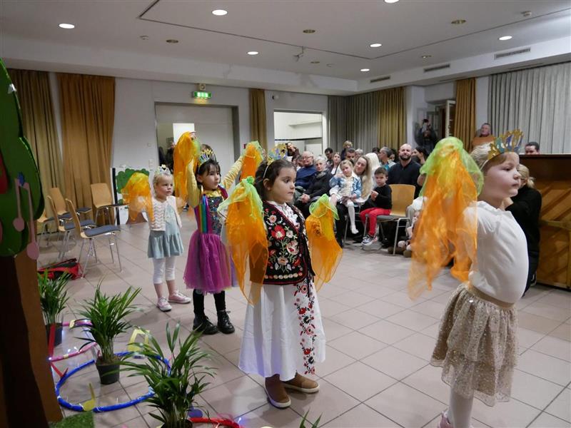 Eine Gruppe von Kindern führt eine Tanz- oder Theateraufführung in einem Gemeinschaftsraum auf. Die Kinder tragen bunte Kostüme mit leuchtenden, orangefarbenen Accessoires und stehen vor einem Publikum aus Erwachsenen und anderen Kindern. Der Raum ist mit Pflanzen und Dekorationen geschmückt, was eine fröhliche und kreative Atmosphäre schafft.