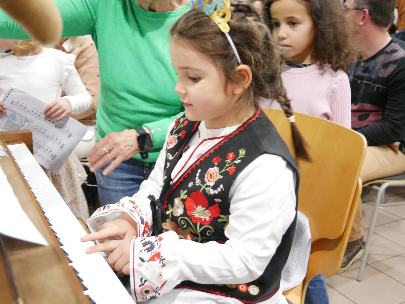 Ein Kind sitzt an einem Klavier und spielt konzentriert. Es trägt eine traditionelle, mit Blumen bestickte Tracht und hat eine goldene Krone im Haar. Im Hintergrund sind weitere Personen zu sehen, darunter ein Kind mit Notenblättern in der Hand.