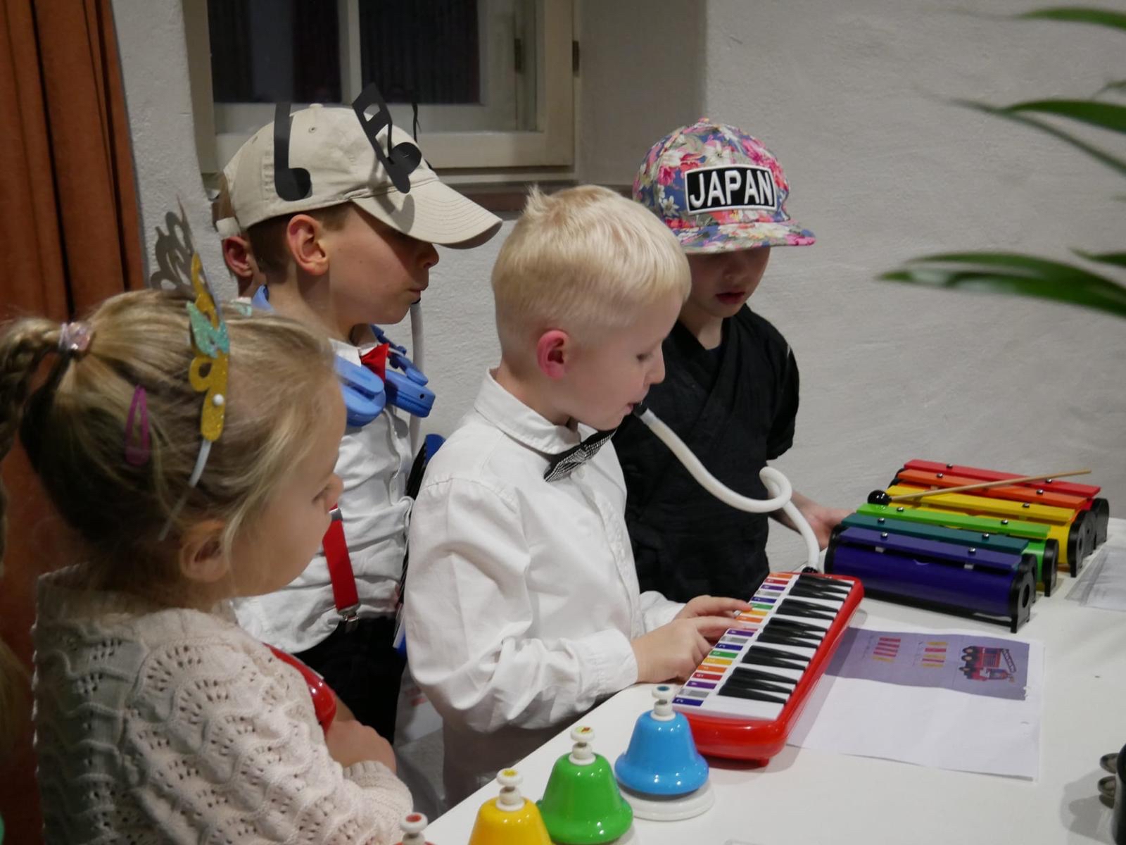 Eine Gruppe von Kindern steht um einen Tisch, auf dem verschiedene Musikinstrumente wie ein buntes Glockenspiel, eine Melodica und farbige Handglocken liegen. Ein Kind spielt auf der Melodica, während die anderen zuschauen. Im Hintergrund ist ein Fenster sowie ein Teil eines Vorhangs und einer Pflanze zu sehen.