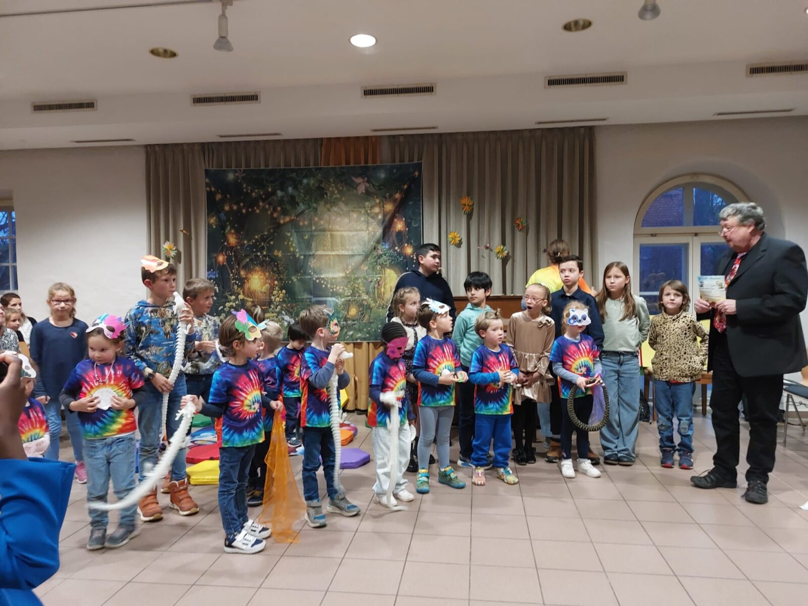 Eine Gruppe von Kindern steht in einem Raum mit dekorativem Hintergrund, der eine märchenhafte Szene mit Lichtern und Pflanzen zeigt. Die Kinder tragen bunte, tie-dye T-Shirts und halten verschiedene Requisiten in der Hand, während sie gemeinsam auf einer Veranstaltung auftreten. Im Hintergrund sind Erwachsene zu sehen, die die Situation beobachten.