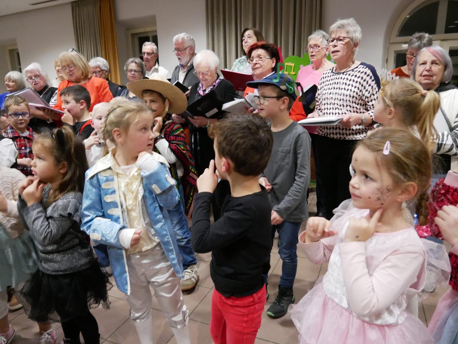 Eine Gruppe von Kindern in bunten Kostümen steht zusammen und scheint an einer Aufführung oder einem gemeinsamen Event teilzunehmen. Im Hintergrund sind Erwachsene mit Notenblättern zu sehen, die möglicherweise ein Lied begleiten. Der Raum ist hell beleuchtet und wirkt wie ein Veranstaltungsraum.