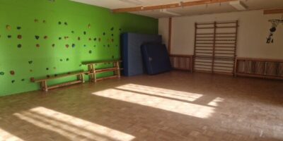 Ein heller Sportraum mit Holzboden und großen Fenstern, durch die Sonnenlicht einfällt. An der grünen Wand befindet sich eine Kletterwand mit bunten Griffen, daneben sind blaue Matten aufgestellt. Rechts ist eine Sprossenwand zu sehen, die für sportliche Aktivitäten genutzt werden kann.
