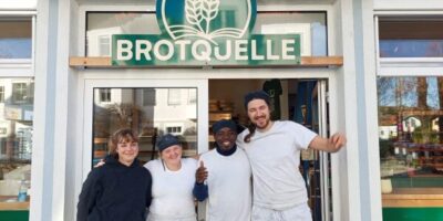 Drei Personen, vermutlich Bäcker, stehen vor der Bäckerei „Brotquelle“ und lächeln in die Kamera. Sie tragen weiße Arbeitskleidung, die Schürzen und T-Shirts kombiniert. Im Hintergrund ist das Schaufenster der Bäckerei mit Produkten zu sehen.