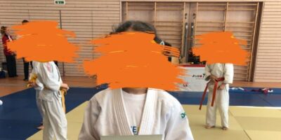 Ein Kind in einem weißen Judoanzug hält stolz eine Urkunde in den Händen. Im Hintergrund sind weitere Kinder in Judoanzügen auf einer Judomatte zu sehen. Die Gesichter der Personen wurden aus Datenschutzgründen unkenntlich gemacht.