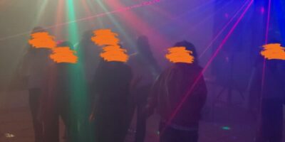 Eine Gruppe von Menschen tanzt in einem Raum mit bunten Lichteffekten und Lasern, die eine lebendige und dynamische Atmosphäre schaffen. Der Raum ist leicht neblig, wodurch die Lichtstrahlen besonders hervorgehoben werden. Die Szene vermittelt das Gefühl einer ausgelassenen Party oder Feier.