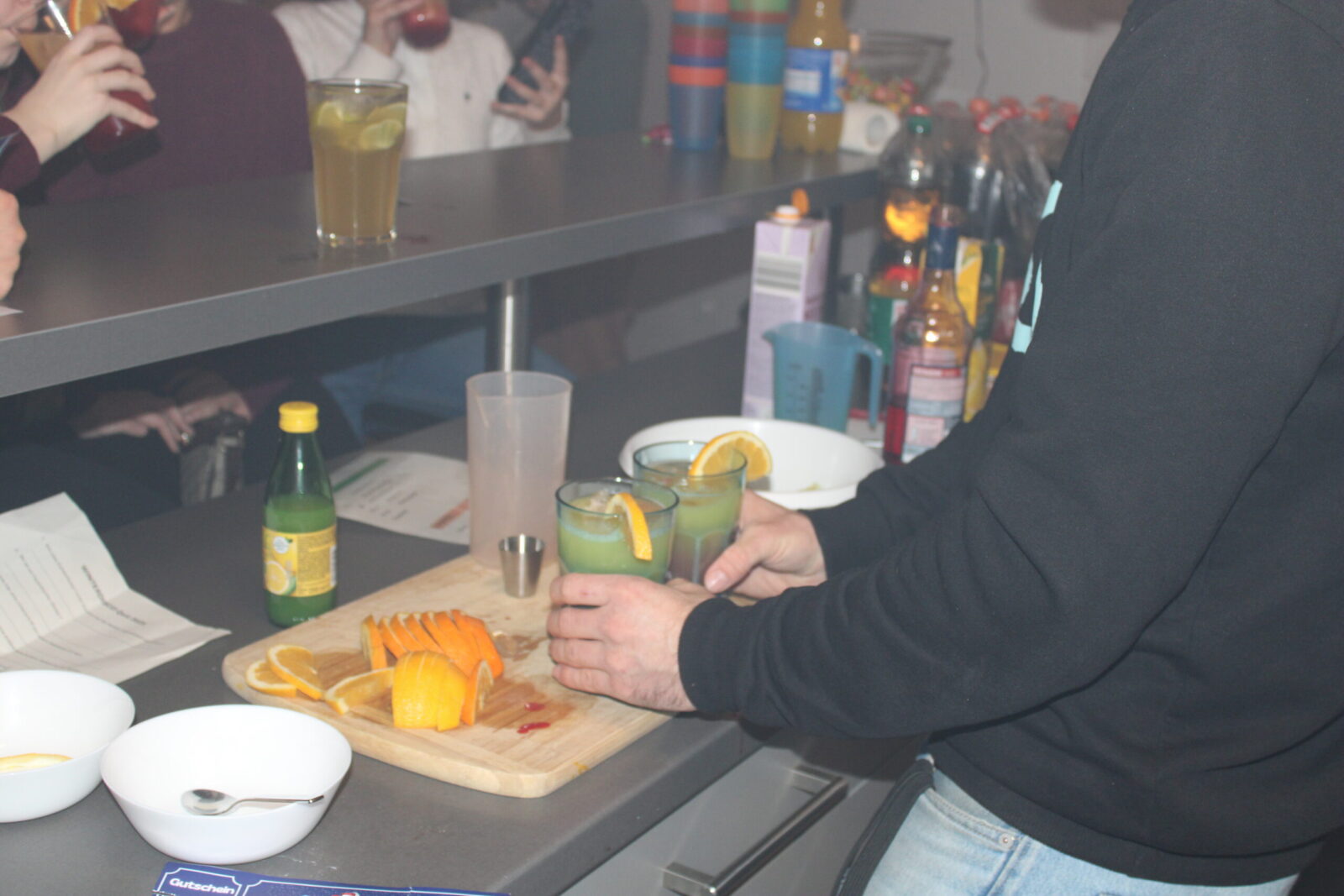 Eine Person bereitet an einer Theke zwei Cocktails mit Orangenscheiben als Garnitur zu. Auf der Theke liegen geschnittene Orangen, eine Zitronensaftflasche und verschiedene Zutaten für Getränke. Im Hintergrund sind weitere Personen zu sehen, die ebenfalls Getränke in der Hand halten.