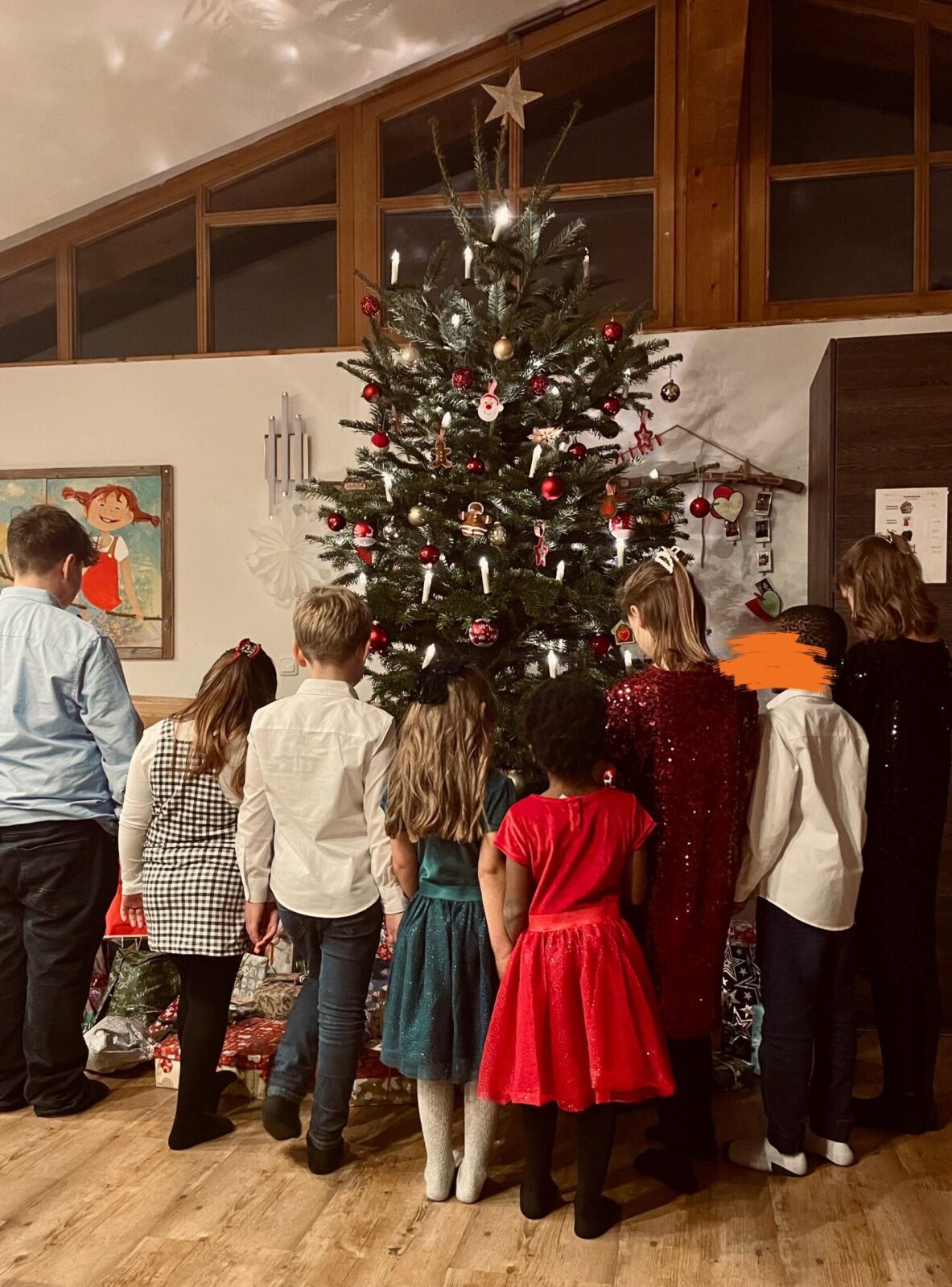 Eine Gruppe von Kindern steht vor einem festlich geschmückten Weihnachtsbaum, der mit roten und goldenen Kugeln sowie Kerzen dekoriert ist. Die Kinder tragen elegante Kleidung in verschiedenen Farben und betrachten die Geschenke unter dem Baum. Im Hintergrund sind ein Fenster und weihnachtliche Dekorationen zu sehen.