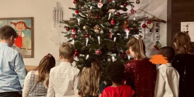 Eine Gruppe von Kindern steht vor einem festlich geschmückten Weihnachtsbaum, der mit roten und goldenen Kugeln sowie Kerzen dekoriert ist. Die Kinder tragen elegante Kleidung in verschiedenen Farben und betrachten die Geschenke unter dem Baum. Im Hintergrund sind ein Fenster und weihnachtliche Dekorationen zu sehen.