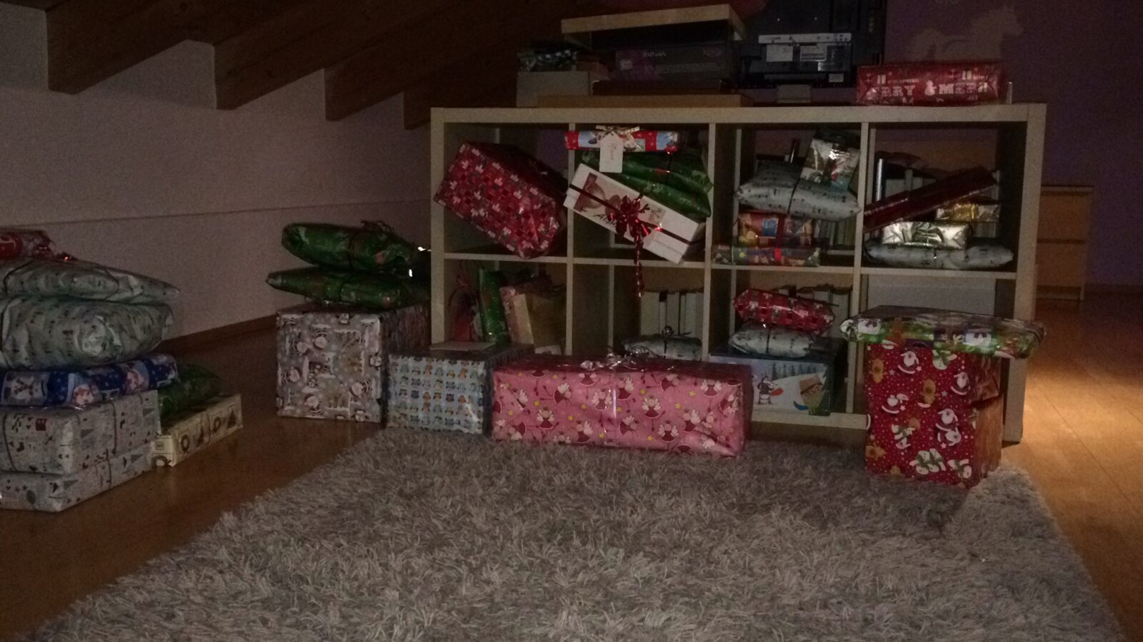 Ein Regal mit mehreren Fächern, in denen zahlreiche bunt verpackte Geschenke liegen. Die Geschenke sind in weihnachtlichem Geschenkpapier gestaltet, darunter rote, grüne und gemusterte Designs. Vor dem Regal liegt ein flauschiger Teppich, auf dem ebenfalls Geschenke platziert sind.