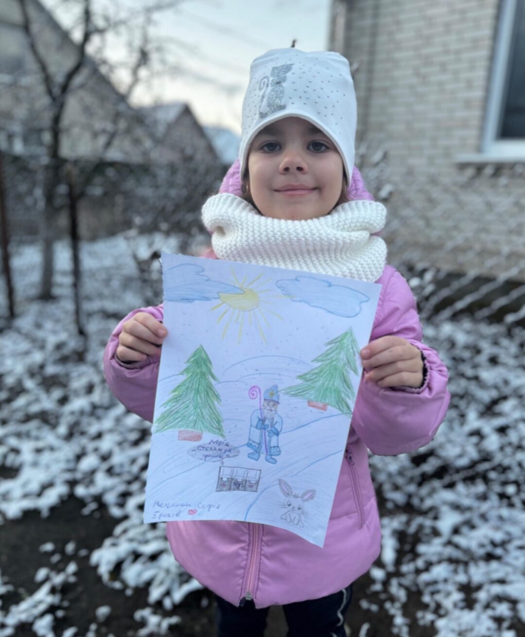 Ein Kind hält eine farbenfrohe Zeichnung in den Händen, die eine Winterlandschaft mit einem Schneemann, zwei Tannenbäumen und einem Hasen zeigt. Im Hintergrund ist eine schneebedeckte Umgebung mit einem Haus und kahlen Bäumen zu sehen. Das Kind trägt eine rosafarbene Jacke und einen weißen Schal, passend zur winterlichen Szenerie.