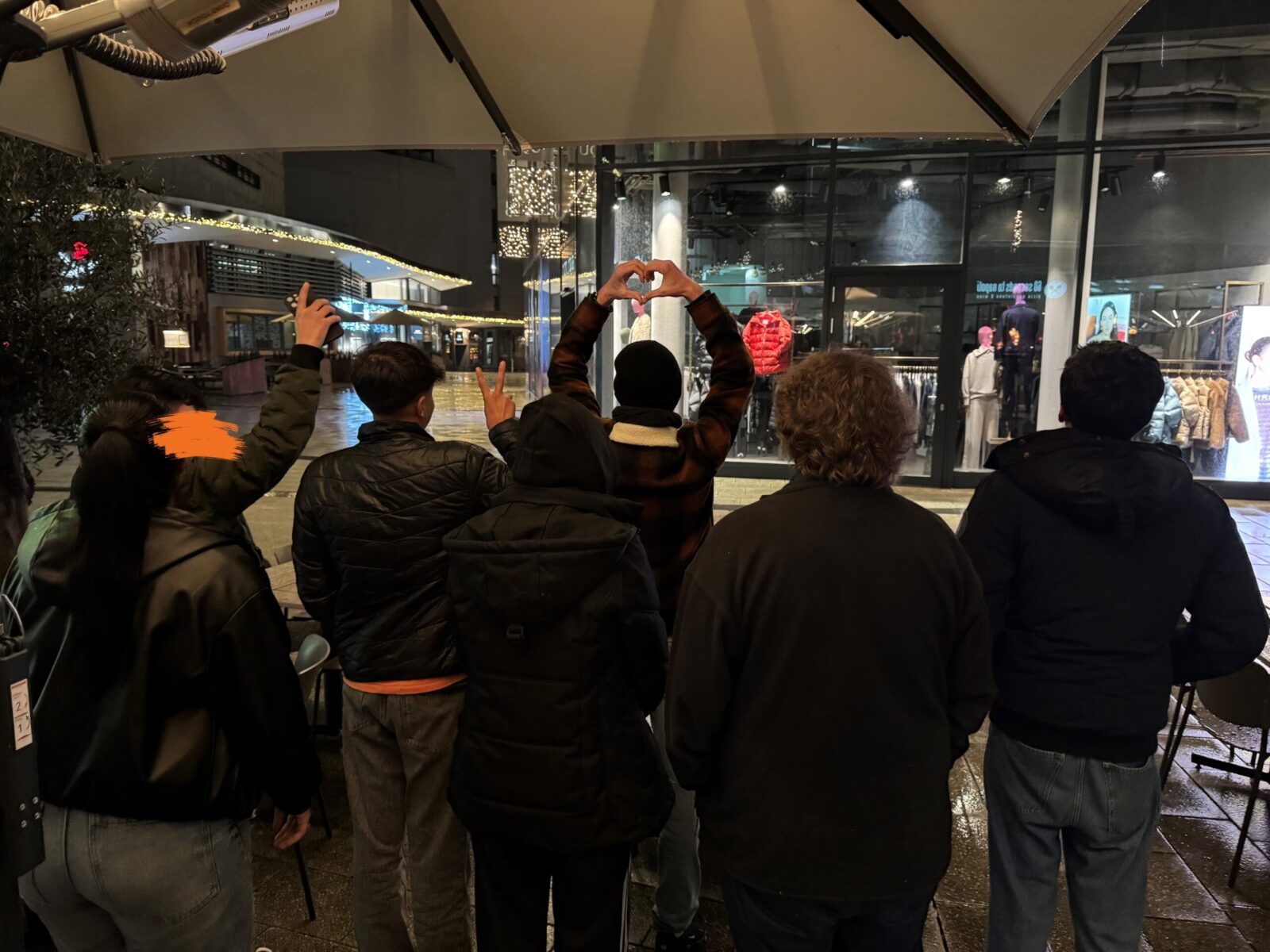 Eine Gruppe von vier Personen steht mit dem Rücken zur Kamera vor einem beleuchteten Schaufenster in einer nächtlichen Stadt. Eine Person formt mit den Händen ein Herz, während eine andere das Peace-Zeichen zeigt. Im Hintergrund sind festliche Lichter und Dekorationen zu sehen, die eine gemütliche Atmosphäre schaffen.